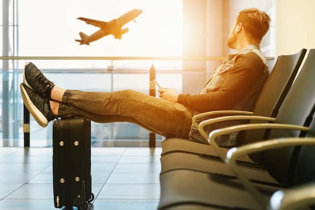 A man setting on gang chair with feet n luggage looking at airplane.