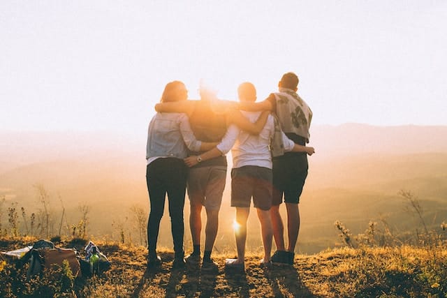 Four people, each one Traveling with Strangers hands wrap around their shoulders while looking at the sunset.