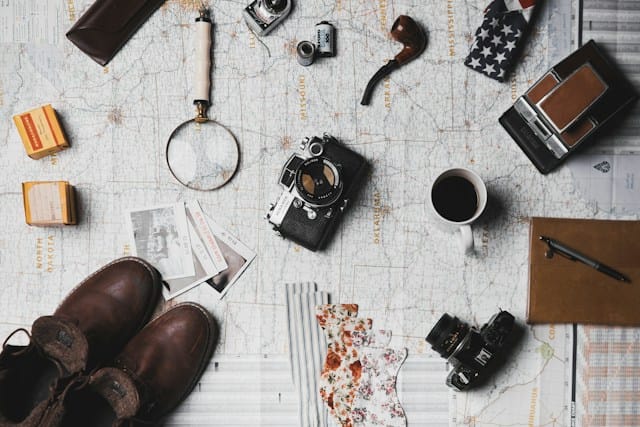  Essential items for travel arrangements: a camera, a pair of brown shoes, a white ceramic mug, grey and black pens, and a brown smoking pipe.